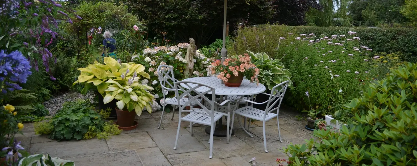 Paved garden patio with a parasol, metal table and chairs surrounded by flower borders